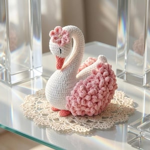 May include: A handcrafted crochet swan figurine. The swan features a white body, a pink beak and feet, and a pink flower on its head. The swan's wings are adorned with fluffy pink yarn. It rests on a cream-colored, crocheted doily.