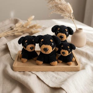 May include: Four handmade crochet dog figurines in black and tan, arranged on a wooden tray. The dogs have detailed features, including button eyes and stitched noses. The background includes a vase with dried plants and a neutral-toned fabric.