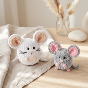 Könnte beinhalten: Zwei gehäkelte Spielzeugmäuse. Eine ist weiß mit beigen Ohren und einer rosa Nase, die andere ist grau mit rosa Ohren und einer rosa Nase. Beide haben schwarze Augen und Schnurrhaare. Sie liegen auf einem beigen Tuch.