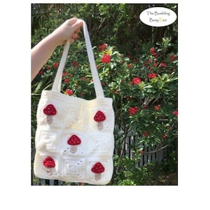 May include: White crocheted tote bag with red and brown mushroom appliques. The bag has two straps and is held by a hand in front of a green bush with red flowers.