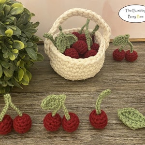 May include: A white crocheted basket filled with red crocheted cherries.  There are also several individual crocheted cherries on a wooden surface.  The text "The Bumbling Busy Bee" is in a yellow box in the upper right corner of the image.