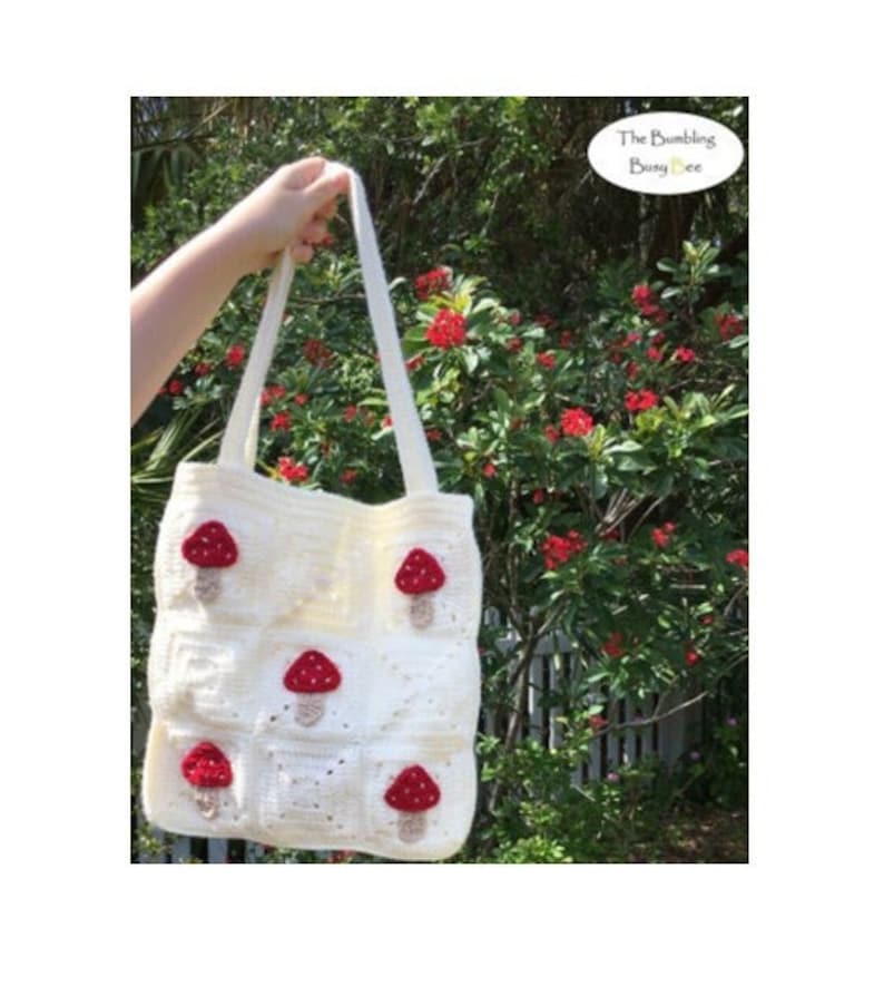 May include: White crocheted tote bag with red and brown mushroom appliques. The bag has two straps and is held by a hand in front of a green bush with red flowers.