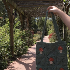 May include: A green crocheted tote bag with mushroom appliques. The bag has a long strap and is held by a hand in the image.