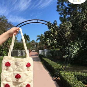 May include: A white crocheted tote bag with red mushroom appliques. The bag has a long strap and is held by a hand in front of a white picket fence and a green hedge.