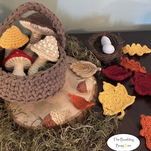May include: A brown crocheted basket filled with crocheted mushrooms in various colors, including red, white, and yellow. The basket is sitting on a wooden slice surrounded by crocheted leaves in shades of red, orange, and yellow. The text "The Bumbling Busy Bee" is visible in the lower right corner of the image.