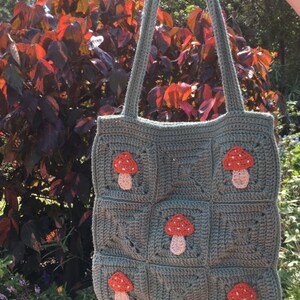May include: A green crocheted tote bag with a mushroom design. The bag has a square shape and features multiple red and pink mushrooms.