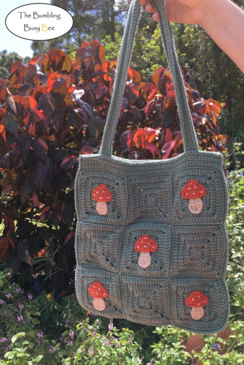 May include: A green crocheted tote bag with a mushroom design. The bag has a square shape and features multiple red and pink mushrooms.