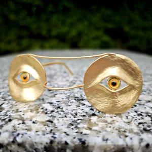 May include: A pair of gold-toned glasses featuring circular frames with a textured, hammered metal finish. Each lens area contains a realistic, detailed eye with a bright orange iris and a black pupil, set directly into the centre of the gold discs.