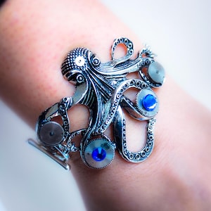 Peut inclure: Un bracelet en argent en forme de pieuvre avec des accents de pierres précieuses bleues et une attache de bracelet de montre.