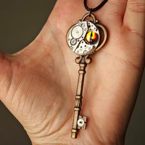 May include: A bronze-toned metal key pendant suspended on a black cord. The bow of the key features a circular watch movement mechanism with exposed silver gears and a central cabochon eye with a yellow iris and a vertical black pupil.