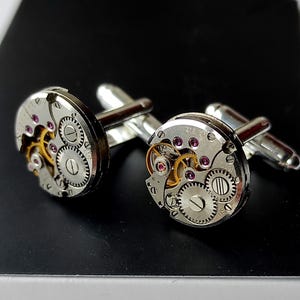 May include: A pair of silver cufflinks with a circular design featuring a detailed watch mechanism with gold gears and pink jewels.