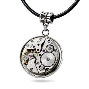 Può includere: Un ciondolo circolare con un movimento di orologio argentato a vista, caratterizzato da ingranaggi, molle e piccole pietre rosse. Il meccanismo è racchiuso in una cornice rotonda argentata, sospesa a un cordino nero tramite una contromaglia lavorata.