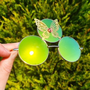 Puede incluir: Gafas de sol redondas con lentes verdes y un detalle de mariposa dorada. La mariposa tiene una gema rosa en el centro. Las gafas de sol tienen una montura plateada. El fondo es un arbusto verde y frondoso.