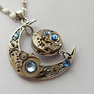 Puede incluir: Un collar con un colgante de media luna de plata con un diseño steampunk. El colgante presenta una esfera de reloj circular con engranajes y cristales azules. El collar está unido a una cadena de plata.