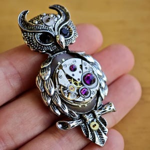 May include: A silver-toned owl brooch featuring a textured metal body, black stone eyes, and a central mechanical watch movement component. The owl's chest is decorated with three round, faceted purple crystals embedded within the clockwork mechanism.