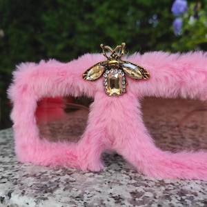 May include: Pink sunglasses with a fluffy, furry texture. The sunglasses have a decorative gold-coloured bee embellishment with clear and amber-coloured crystals. The frames are a vibrant pink.