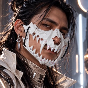 Op de afbeelding: Een wit, getextureerd gezichtsmasker met een grillig, tandachtig ontwerp langs de onderrand, geaccentueerd met kleine, iriserende en barnsteenkleurige kristallen. Het masker is bevestigd met zwarte banden over de onderkant van het gezicht.