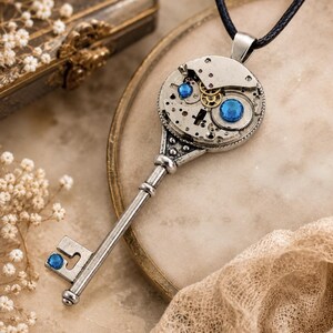 Può includere: Una collana con ciondolo a forma di chiave color argento dotata di un meccanismo a movimento d'orologio circolare sulla parte superiore. L'assemblaggio meccanico include ingranaggi in ottone a vista e due gemme circolari blu brillante incastonate.