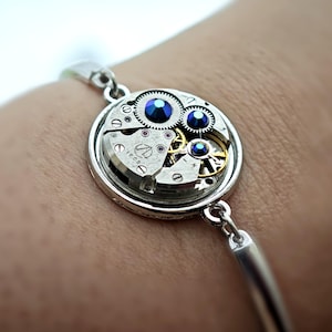 May include: Silver-toned bracelet featuring a watch movement centerpiece. The round pendant showcases intricate gears, jewels, and a small inscription. Two blue gems add a pop of color. The bracelet is attached to a slender, curved band.