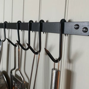 May include: Black metal pot rack with hooks, mounted on a white wall.  Various kitchen utensils are hanging from the hooks.