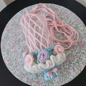 May include: A handmade crochet phone bag in a light pink net design with a matching strap. The bottom of the bag is decorated with blue, white, pink, and purple crochet flowers and a small bow. The bag is on a floral patterned surface.