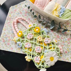 Puede incluir: Un bolso de ganchillo hecho a mano con un diseño de red verde, adornado con flores de ganchillo coloridas en amarillo, rosa y azul. El bolso tiene un asa rosa y se apoya sobre una tela con estampado floral. Una cesta de hilo en varios colores pastel está en el fondo.