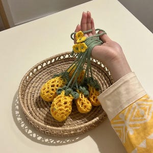 May include: Crocheted yellow pineapple ornaments with green stems and yellow flowers are held above a woven basket. The pineapples have a textured, open weave design. The background is a light-colored table.