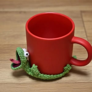 May include: A red mug with a green crocheted lizard wrapped around it. The lizard has white eyes and a pink tongue sticking out. The mug is sitting on a wooden surface.
