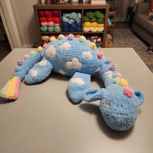 May include: A plush blue dinosaur-shaped pillow with white cloud appliqués and colorful pom-poms. The dinosaur has a rainbow-colored tail and is made of soft, fuzzy material. A whimsical and cuddly decorative item.