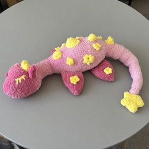May include: A handmade pink and yellow crochet Nessie plush toy. The plush has a long neck and tail, with yellow flower-shaped accents on its back and head. The toy is lying on a gray surface.