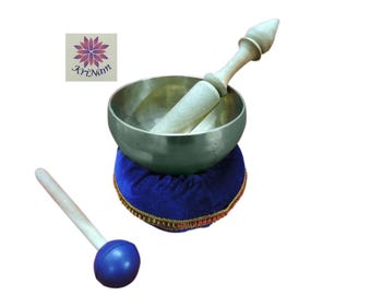May include: A silver-colored singing bowl with a wooden mallet, resting on a blue velvet cushion with gold trim. A second mallet with a blue head is also present. The logo "KriNam" is in the upper left corner.
