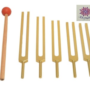 May include: A set of gold-coloured tuning forks and a wooden mallet with a red head. The tuning forks are arranged in a row. A small square image with a floral design and text is in the upper right corner.