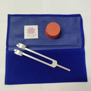 May include: A silver tuning fork with a red handle, resting on a blue velvet pouch. An orange cylinder and a sticker with the text "Kri Nam" are also visible. The tuning fork is designed for sound healing and meditation.