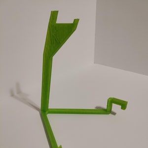 May include: A green 3D printed stand with three legs and a curved top. The stand is designed to hold a small object, such as a phone or a tablet.