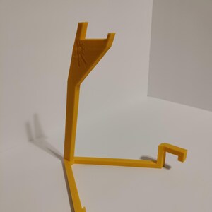 May include: A yellow plastic stand with three legs and a curved top. The stand is designed to hold a small object, such as a phone or a tablet.