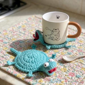May include: A cream-colored mug with a brown handle and a cat illustration sits on a blue crocheted frog coaster. Another frog coaster and a floral patterned placemat are also visible. A white spoon rests on the placemat.