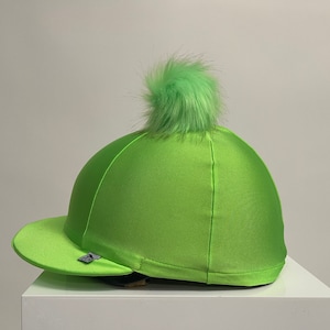 May include: A vibrant green riding helmet cover with a peak and a fluffy pom-pom. The cover is made from a smooth, satin-like fabric and has a small logo. The helmet is displayed on a white surface, showcasing its bright colour.