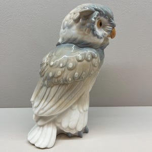 K&ouml;nnte beinhalten: Eine Eulenfigur aus Porzellan mit gl&auml;nzender Oberfl&auml;che in den Farben sanftes Grau, Beige und Wei&szlig;. Die Eule ist im Profil dargestellt und zeigt detaillierte Federmuster, ein kleines orangefarbenes Auge und einen hellbraunen Schnabel.