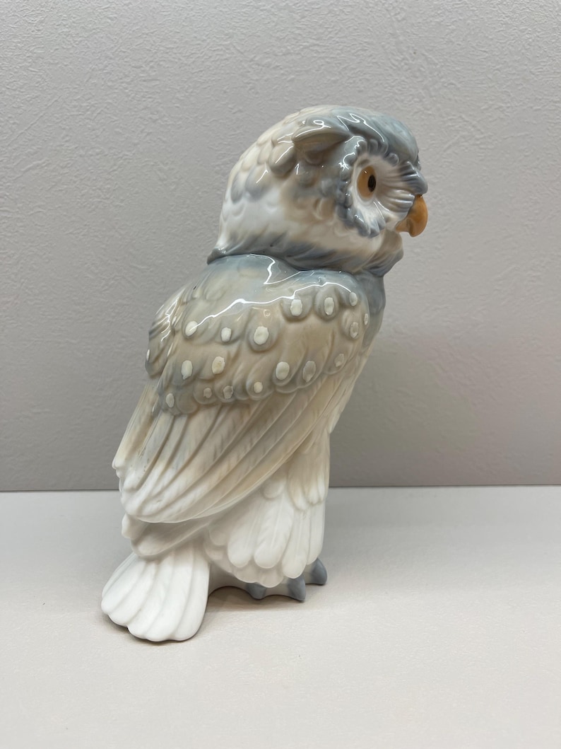 K&ouml;nnte beinhalten: Eine Eulenfigur aus Porzellan mit gl&auml;nzender Oberfl&auml;che in den Farben sanftes Grau, Beige und Wei&szlig;. Die Eule ist im Profil dargestellt und zeigt detaillierte Federmuster, ein kleines orangefarbenes Auge und einen hellbraunen Schnabel.
