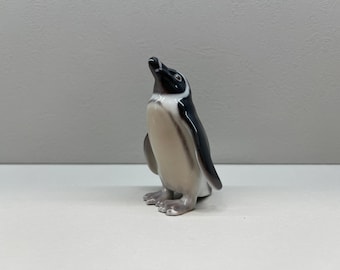 May include: A small porcelain penguin figurine with black and white markings. The penguin is standing upright with its head tilted upwards.