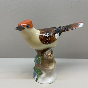 Puede incluir: Una figurita de porcelana de un pájaro con la cabeza roja, alas marrones y blancas y cola marrón. El pájaro está posado en un pequeño tocón de árbol con hojas verdes.