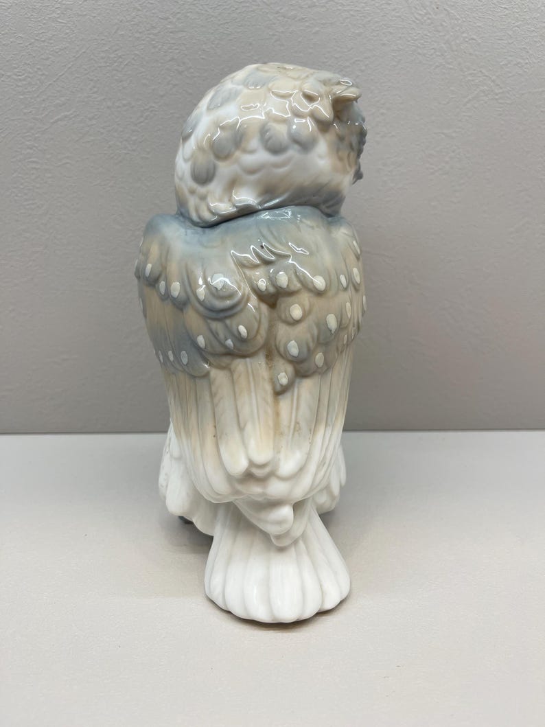 K&ouml;nnte beinhalten: Eine Eulenfigur aus Porzellan mit gl&auml;nzender Oberfl&auml;che in einer Farbpalette aus sanftem Grau, ged&auml;mpftem Blau und Cremewei&szlig;. Die Eule ist von hinten dargestellt und zeigt detaillierte Federstrukturen sowie geschichtete Fl&uuml;gelmuster.