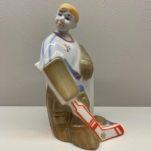 May include: A ceramic figurine of a hockey player in a white and blue striped jersey, brown pants, and a hockey stick. The figurine has a blonde head and is in a kneeling position. The hockey stick is white with orange stripes and a snowflake design.