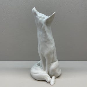 Puede incluir: Una figurita de porcelana blanca de un zorro sentado con la cabeza levantada. El zorro está detallado con pelaje y tiene una cola larga y tupida.