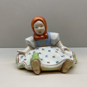 Puede incluir: Una figurita de porcelana de una niña que lleva un vestido rojo y azul con un estampado floral blanco. Está sentada con las piernas cruzadas y tiene una expresión de sorpresa en su rostro.