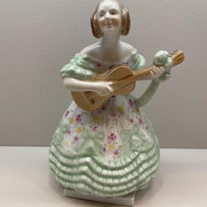 Puede incluir: Una figurita de porcelana de una mujer tocando la guitarra. Lleva un vestido verde y blanco con detalles florales y una guitarra dorada.