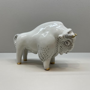 May include: White ceramic figurine of a bison with gold accents. The bison is standing with its head turned to the side.