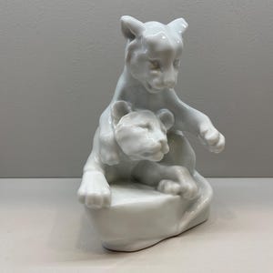 Puede incluir: Una figurita de porcelana blanca de dos cachorros de león jugando. El cachorro más grande está de pie sobre sus patas traseras, mientras que el más pequeño está sentado en una roca. La figurita es un ejemplo clásico de escultura animal.