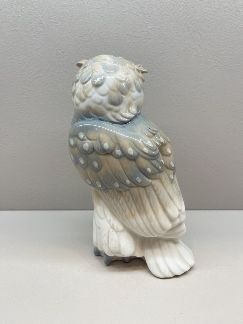 K&ouml;nnte beinhalten: Eine Eulenfigur aus Porzellan in einer sanften Farbpalette aus ged&auml;mpftem Blau, Beige und Wei&szlig;. Die Eule ist im Seitenprofil dargestellt und zeigt detaillierte Federstrukturen, kleine wei&szlig;e Punkte auf den Fl&uuml;geln und eine glatte Oberfl&auml;che.
