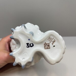 K&ouml;nnte beinhalten: Die Unterseite einer wei&szlig;en Porzellanfigur mit gl&auml;nzender Oberfl&auml;che und bl&auml;ulich get&ouml;nten Kanten. Der Boden zeigt die handgemalte schwarze Zahl 30, ein braunes quadratisches Herstellersiegel sowie den Text DAISA, LLADRO und handmade in Spain.